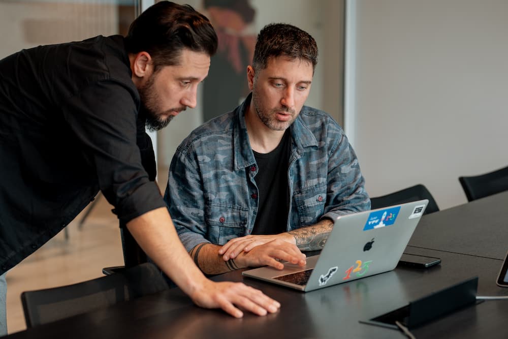 Dois homens colaboram em uma laptop prateada. Um está sentado e o outro de pé, inclinado sobre a mesa, observando a tela.