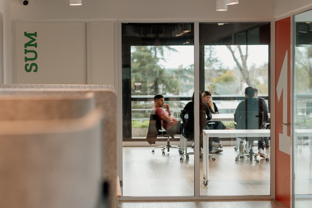 Uma vista através de uma parede de cristal para uma sala de reuniões onde se vêem pessoas sentadas ao redor de uma mesa. No primeiro plano, se vê parte de um letrero verde com a palavra 'SUM' em uma parede.