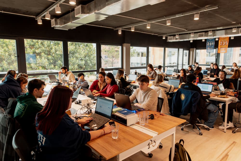 Uma oficina grande e moderna com várias pessoas trabalhando em seus laptops em escritórios de madeira. Grandes janelas oferecem vistas para o exterior.
