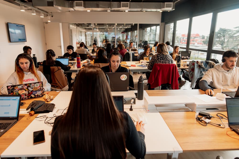 Uma vista desde detrás de uma mulher em uma oficina aberta, mostrando a várias pessoas trabalhando em seus laptops. O espaço é amplo com grandes janelas no fundo.