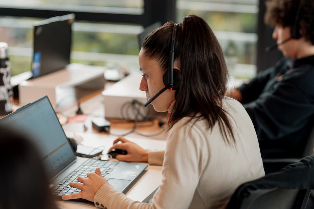 Uma mulher com fones de ouvido e microfone trabalha em sua laptop em uma oficina. Está concentrada na tela, com sua mão no rato e a outra no teclado.