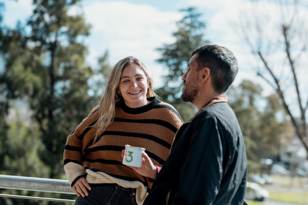 Um homem e uma mulher se divertem conversando ao ar livre, possivelmente em uma terraza. A mulher usa um suéter a rayas e o homem segura uma xícara com o número '3'. Há árvores e céu azul no fundo.