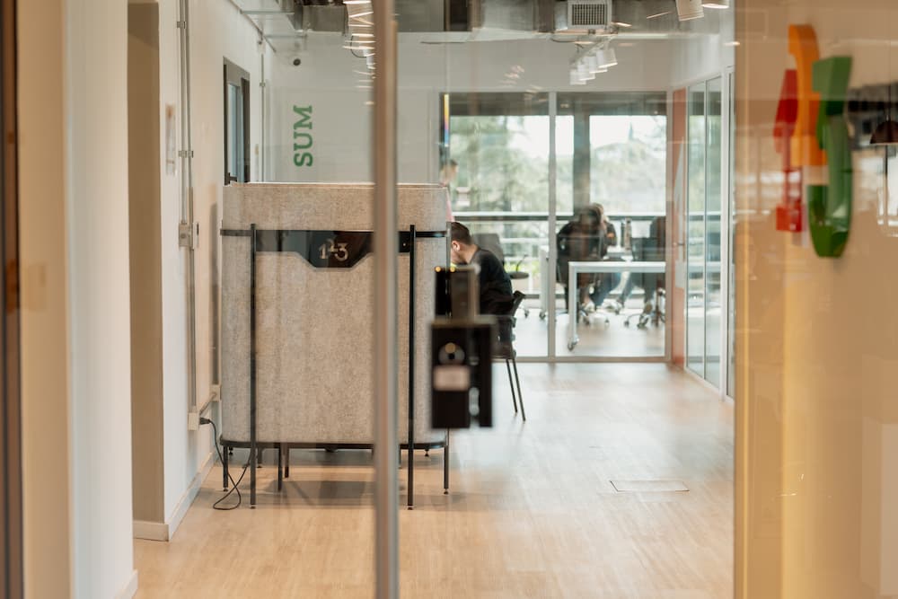 Uma vista através de uma parede de cristal em uma oficina moderna. Se vê um espaço de trabalho com uma pessoa sentada no fundo, e no primeiro plano, divisores de escritório com um letrero que diz 'SUM' na parede.
