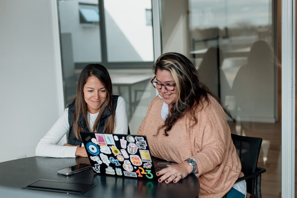 Duas mulheres olham uma laptop cheia de adesivos em uma oficina. A mulher da direita, que usa óculos, sorri e aponta para a tela.