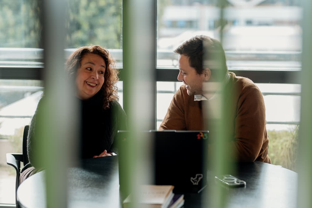 Um homem e uma mulher se divertem conversando, sentados em frente a uma laptop. A imagem está ligeiramente desfocada por elementos no primeiro plano.