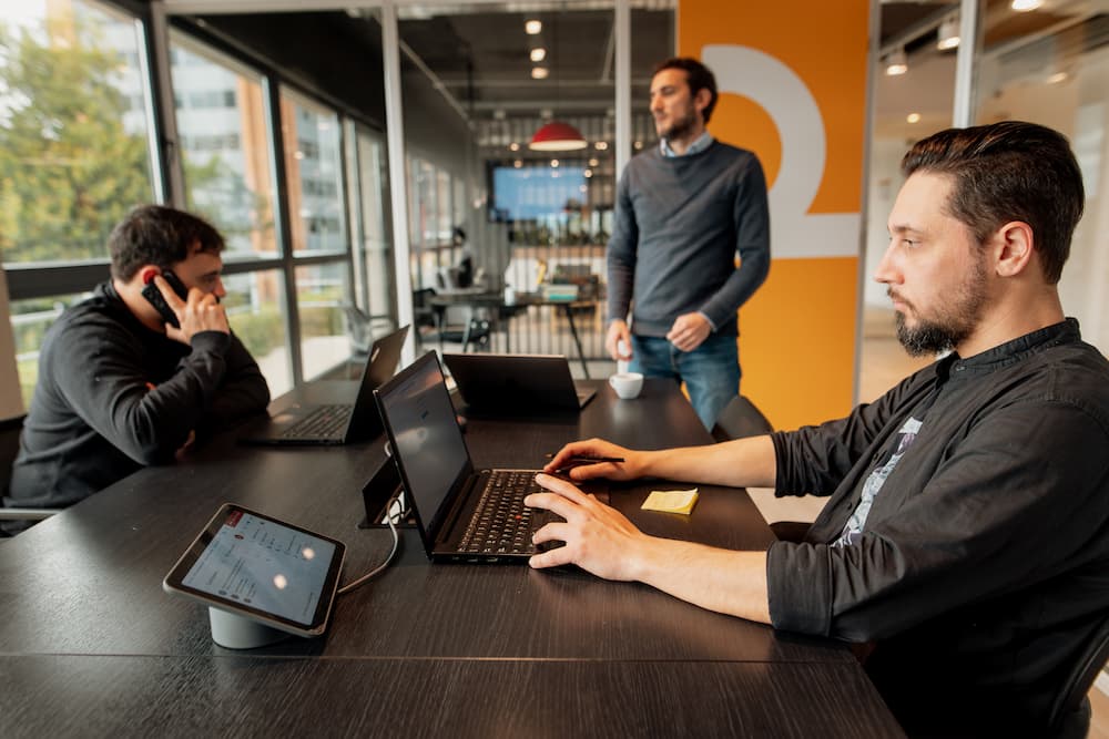 Três homens trabalham em uma oficina moderna. Um está no telefone, outro tecleia em sua laptop e o terceiro está de pé, olhando para um lado. Há uma tablet com uma base no primeiro plano.