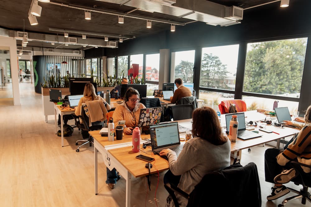 Um plano geral de uma oficina moderna e luminosa com várias pessoas trabalhando em escritórios individuais com laptops. Grandes janelas oferecem vistas para o exterior.
