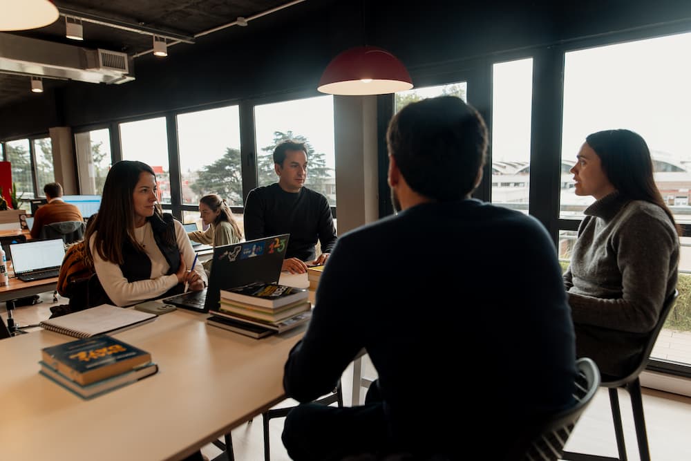 Um grupo de quatro pessoas conversa ao redor de uma mesa em uma oficina aberta. Há laptops e livros sobre a mesa, e grandes janelas no fundo.